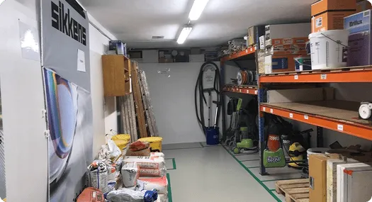 Lagerraum mit Stapeln von Baumaterialien und Reinigungsgeräten auf Regalen und Boden.
