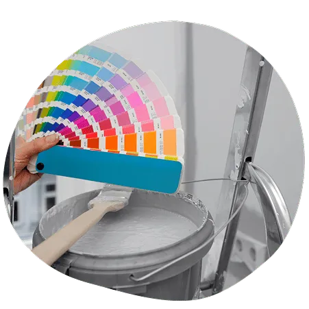 Hand holding a color swatch fan above a bucket of gray paint with a paintbrush.