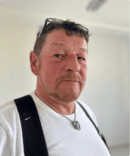 Porträt eines Mannes mit Brille auf dem Kopf, weißem T-Shirt und Halskette vor unscharfem Hintergrund.