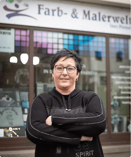 Frau mit kurzen schwarzen Haaren und Brille steht vor einem Geschäft mit dem Schild 'Farb- & Malerwelt'.