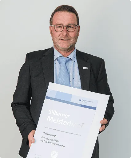 Mann in Anzug und blauer Krawatte hält ein Silberner Meisterbrief Zertifikat vor weißem Hintergrund.