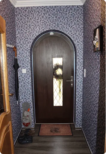 Eingangsbereich mit dunkler Holztür, Verzierung aus Glas mittig, blau-gemusterter Tapete und einem Türvorleger.
