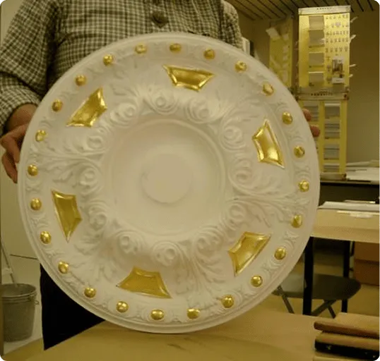 Person hält eine verzierte, runde weiße Deckenrosette mit goldenen Akzenten.