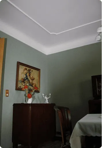 Wohnzimmer mit grün gestrichener Wand, einem Holzschrank mit Blumenstrauß, einem Stuhl und einem gedeckten Tisch mit weißer Tischdecke.