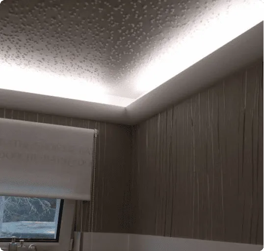 Decke mit indirekter Beleuchtung in einem Raum mit einer aufgerollten weißen Rolloscheibe und einem Fenster mit Bergblick.