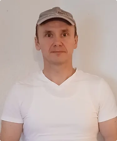 Mann mit beiger Kappe und weißem T-Shirt vor einer hellen Wand.