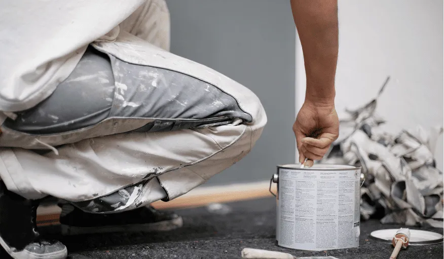 Ein Handwerker in Arbeitskleidung hält einen Pinsel über eine Farbdose auf einem Boden mit Malerfolie.